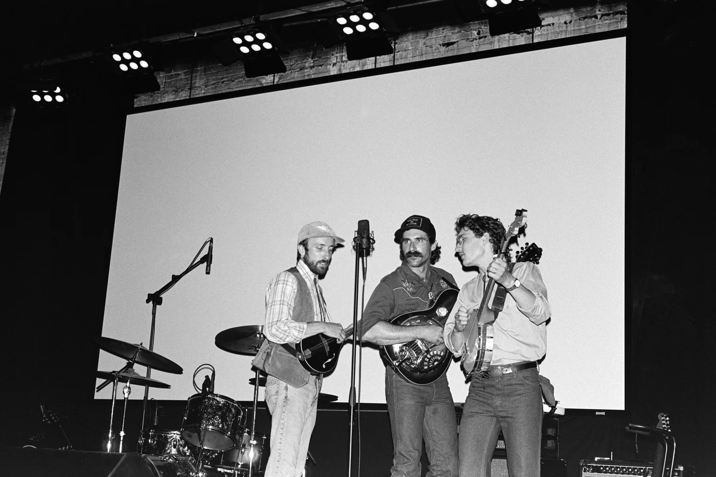three country folk singers around a single microphone on a stage with professional lighting.